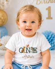 A smiling baby boy wearing a white t-shirt with a personalized name and animal print, sitting in front of a birthday party backdrop with balloons and confetti.
