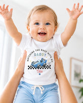 A smiling baby wearing a white t-shirt with a racing car graphic and the text "WATCHING THE RACE WITH MY DADY" is being held up by someone's arms.