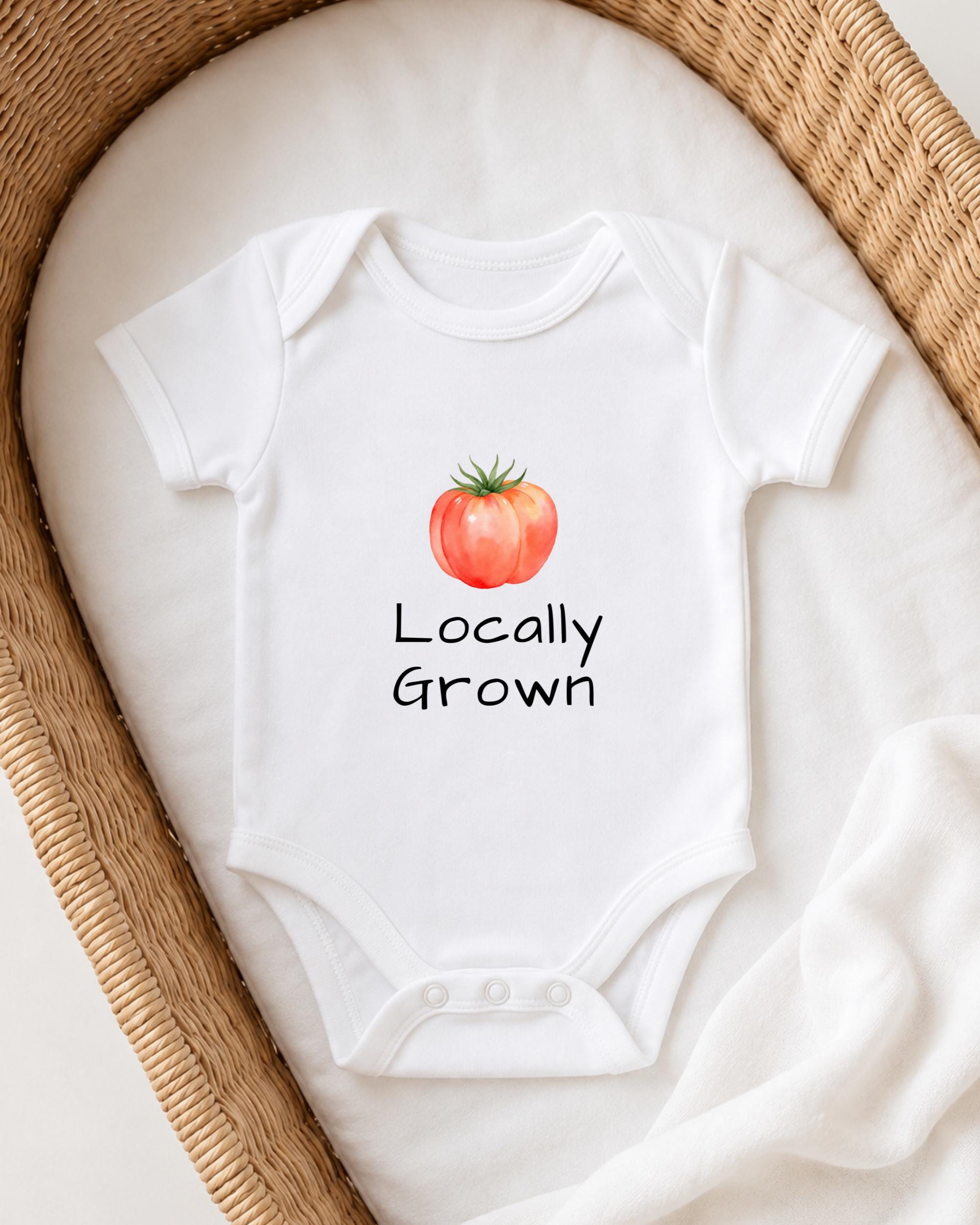 A white baby onesie with a tomato graphic and the text "Locally Grown" printed on it, placed in a woven basket.