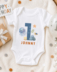 A white onesie with a blue number 1 and various marine life designs, including an octopus, starfish, and sea turtle, is displayed on a white blanket.