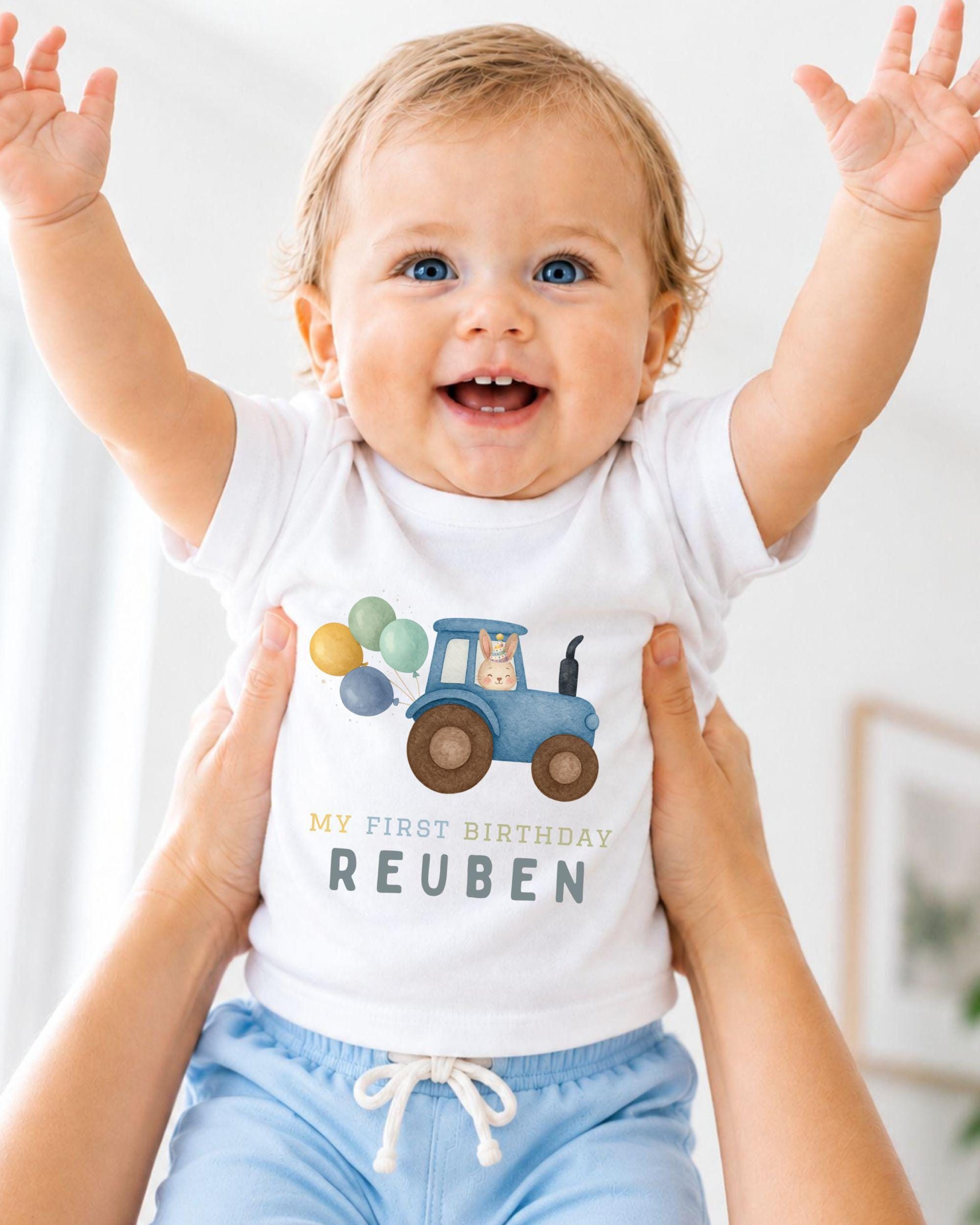 A smiling baby wearing a white t-shirt with a tractor graphic and the text "My first birthday Reuben" is being held up by someone's arms.