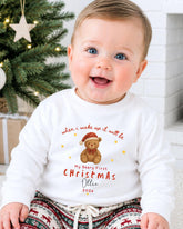 A smiling baby wearing a white sweater with a teddy bear and text that says "When I wake up it will be my first Christmas, Ollie 202x".