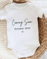A white baby onesie with the text "Coming Soon October 2025" printed on it, placed on a woven basket.