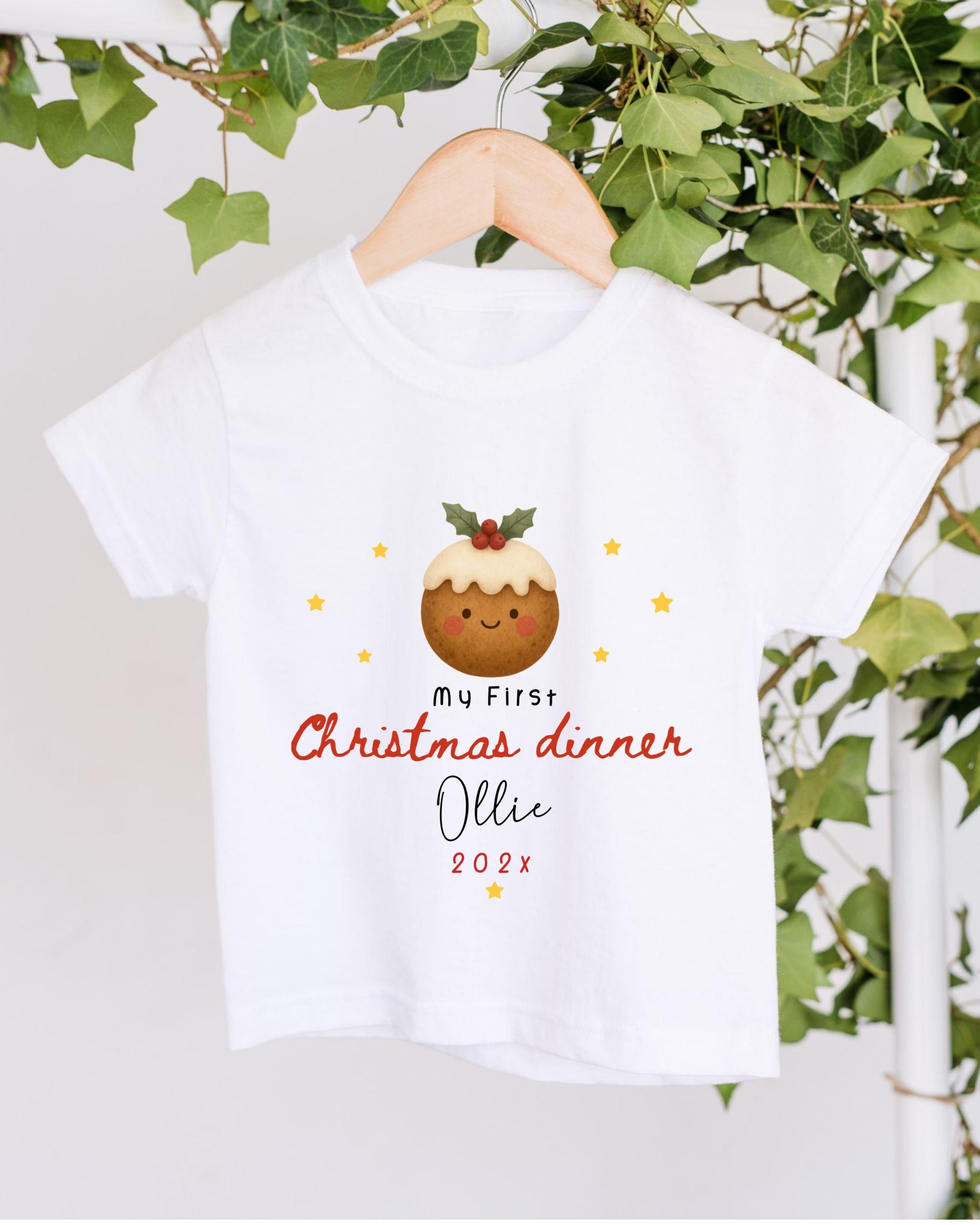 A white t-shirt with a Christmas-themed design hanging on a wooden hanger, surrounded by greenery.