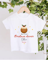 A white t-shirt with a Christmas-themed design hanging on a wooden hanger, surrounded by greenery.