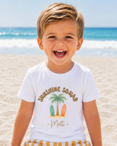 A young boy with a big smile on his face, wearing a white t-shirt with a graphic design of a palm tree and surfboards, standing on a sandy beach with the ocean in the background.