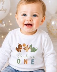 A smiling baby boy wearing a white shirt with a dinosaur and a bird graphic, and the text "Jack is one" printed on it.