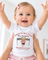 A smiling baby girl wearing a white t-shirt with a teddy bear and the text "Cheering for My Mummy" is being held up by someone's arms.