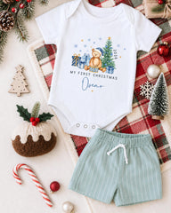 A white onesie with a teddy bear and Christmas decorations, a pair of green shorts, and other holiday-themed items are arranged on a plaid blanket.