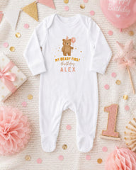 A white onesie with a teddy bear and the text "My first birthday Alex" printed on it, surrounded by pink and gold confetti, a pink balloon, and other baby-related items.