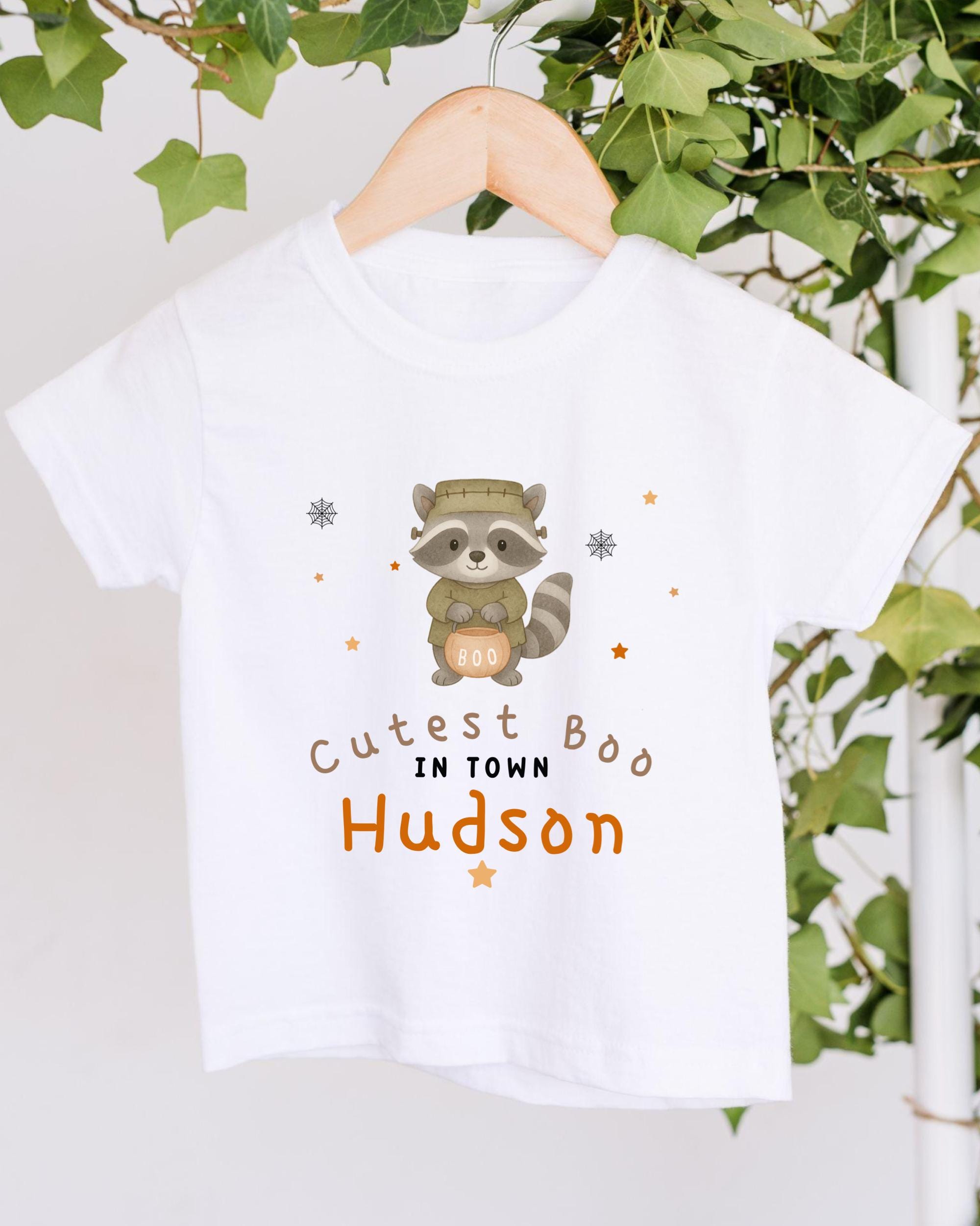 A white t-shirt with a raccoon graphic and text that says "Cute in town Hudson" hanging on a wooden hanger.