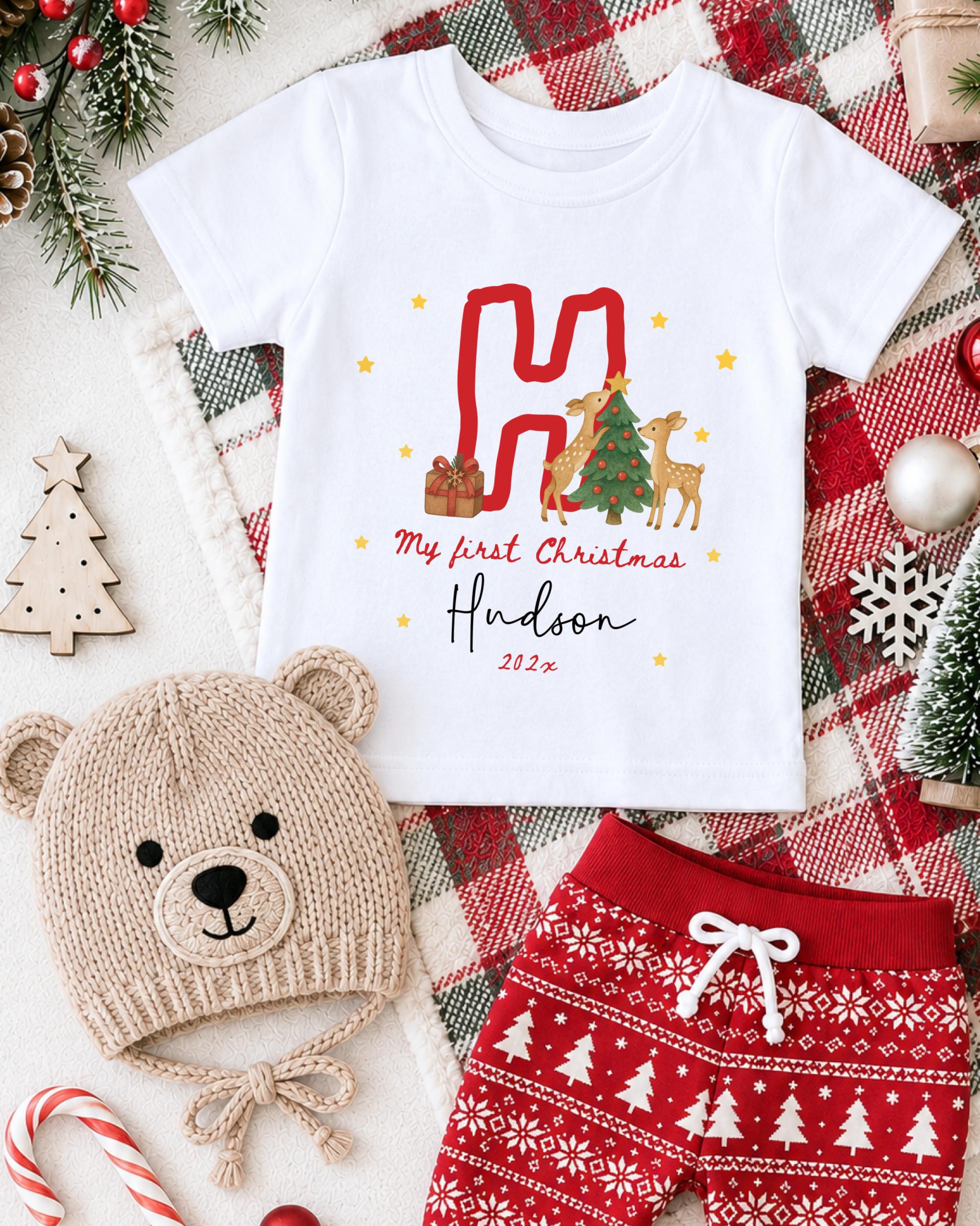 A white t-shirt with a Christmas-themed design, a beige knit hat, red shorts, and other Christmas-themed items are arranged on a plaid blanket.