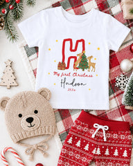 A white t-shirt with a Christmas-themed design, a beige knit hat, red shorts, and other Christmas-themed items are arranged on a plaid blanket.