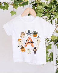 A white t-shirt with a graphic design featuring various cartoon-style characters, including a bat, skeleton, ghost, pumpkin, and a letter "A" surrounded by stars.