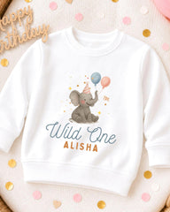 A white sweatshirt with a graphic of an elephant wearing a party hat and holding balloons, along with the text "Wild One ALISHA".