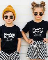 Two children wearing black t-shirts with the text "BOO Jackie" and round sunglasses, standing against a white wall.