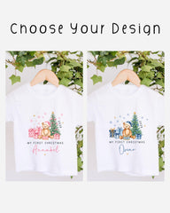 two white t-shirts with Christmas-themed designs, hanging on a wooden hanger. The t-shirts feature illustrations of a bear, a Christmas tree, and presents, along with the text "My first Christmas" and "My first Christmas" written on them.