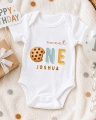 A white onesie with the text "Sweet One Joshua" printed on it, along with a cookie icon, is displayed on a bed with a "Happy Birthday" sign and a balloon.