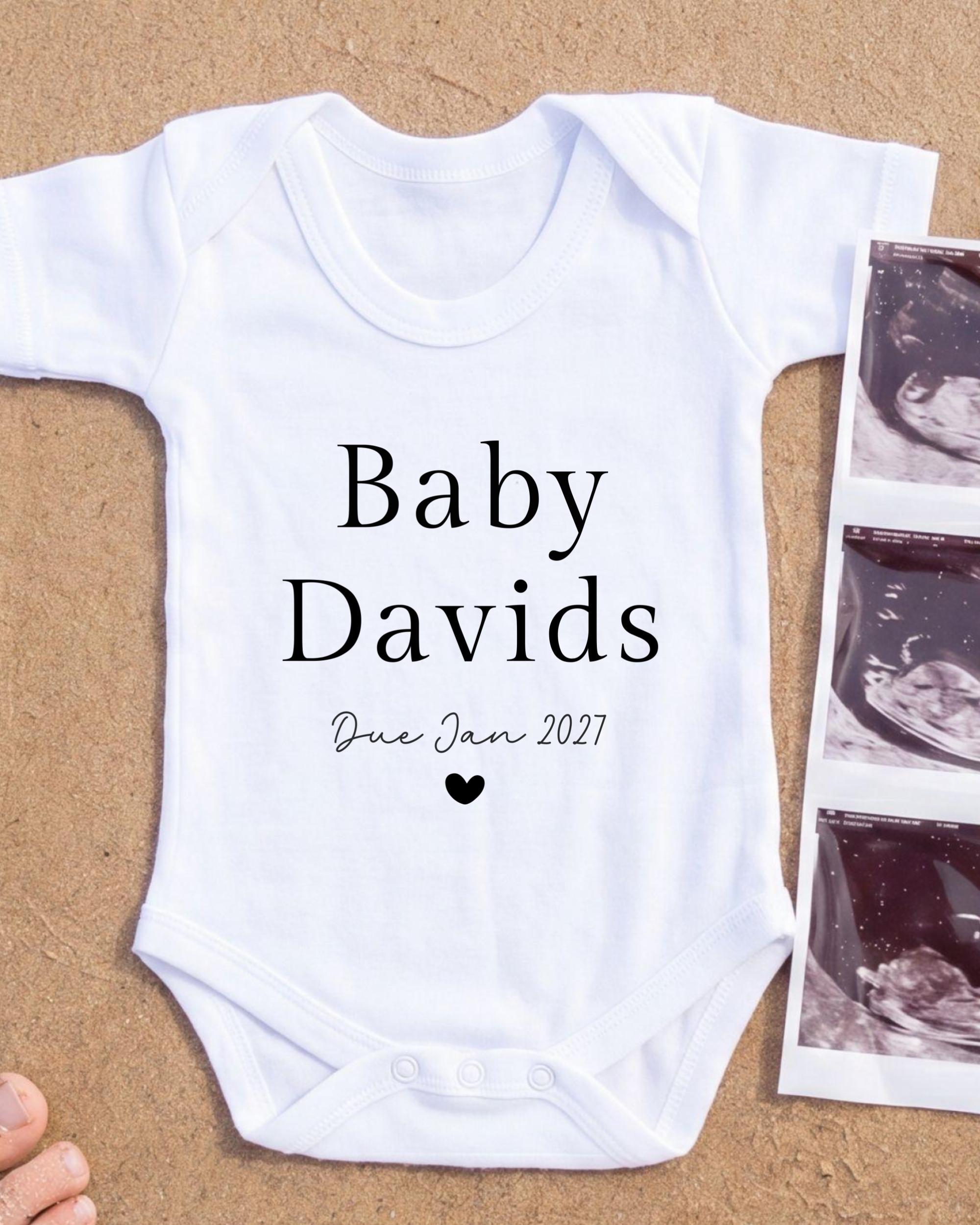 A white baby onesie with the text "Baby Davids Due Jan 2027" printed on it, along with a heart symbol. The onesie is placed next to a photograph of an ultrasound image.