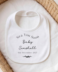 A white bib with black text that reads "Sea You Soon Baby Seashell Due December 2027".