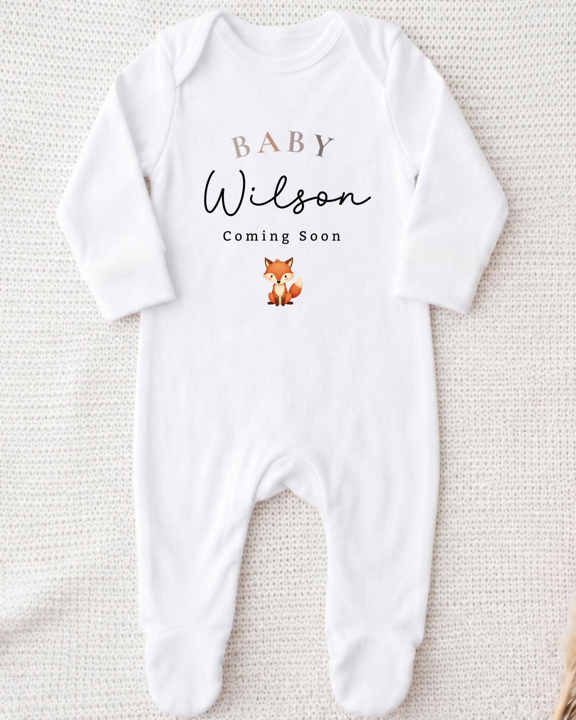 A white onesie with the text "BABY WILSON Coming Soon" printed on it, along with a small illustration of a fox.