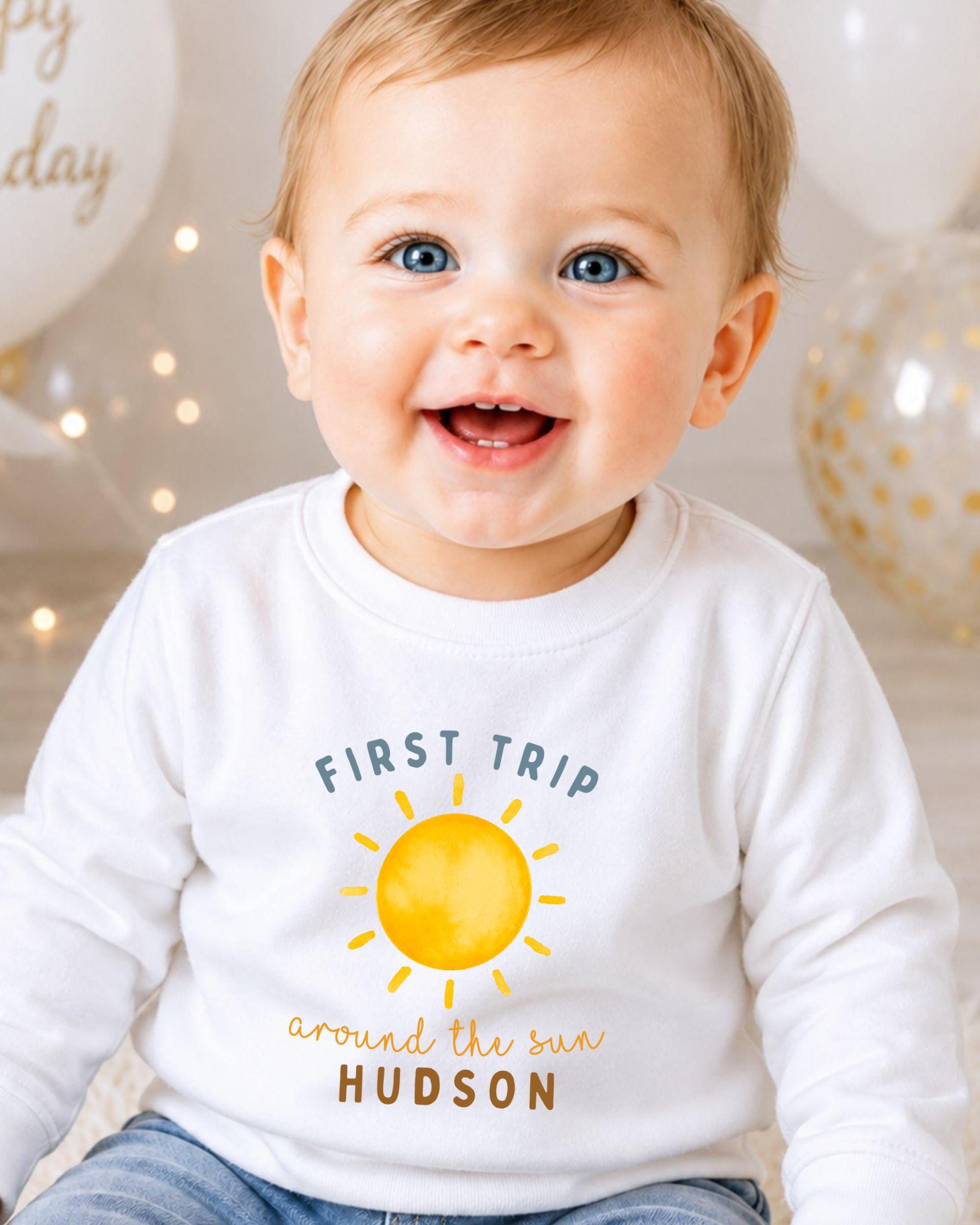 A smiling baby boy wearing a white shirt with a sun graphic and text that says "First Trip around the Sun Hudson".