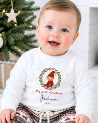 A smiling baby boy wearing a white sweater with a Christmas-themed design and text, sitting in front of a Christmas tree.
