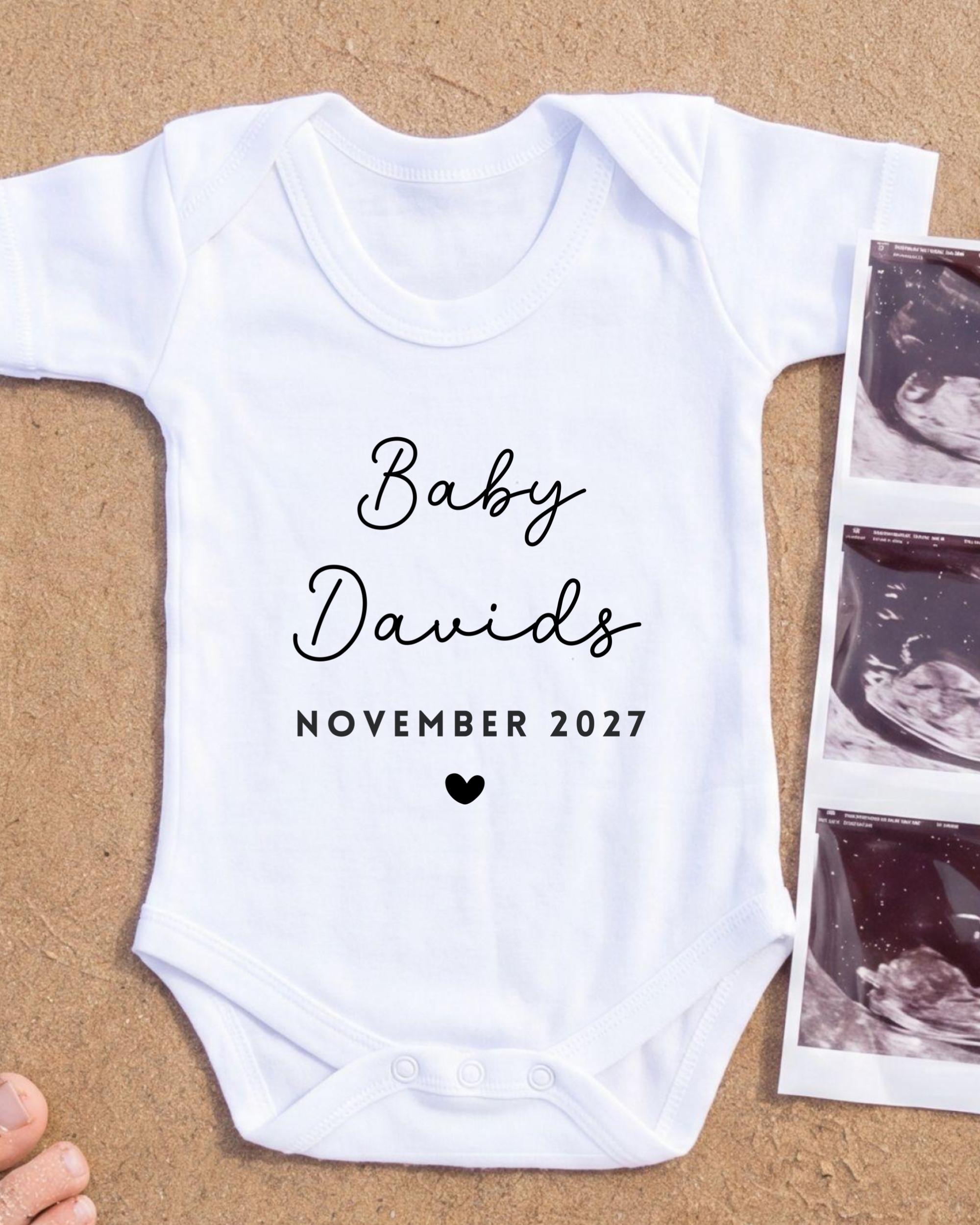 A white baby onesie with the text "Baby Davids November 2027" printed on it, along with a heart symbol. The onesie is placed next to a photograph of an ultrasound image.