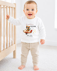 A smiling baby boy wearing a white sweater with a cartoon airplane and the text "First Time Flyer Mike" is standing in a nursery.