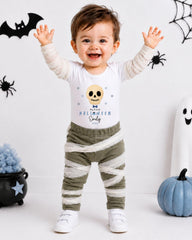 A young child wearing a Halloween-themed shirt and striped pants stands with their arms raised, smiling and surrounded by Halloween decorations.