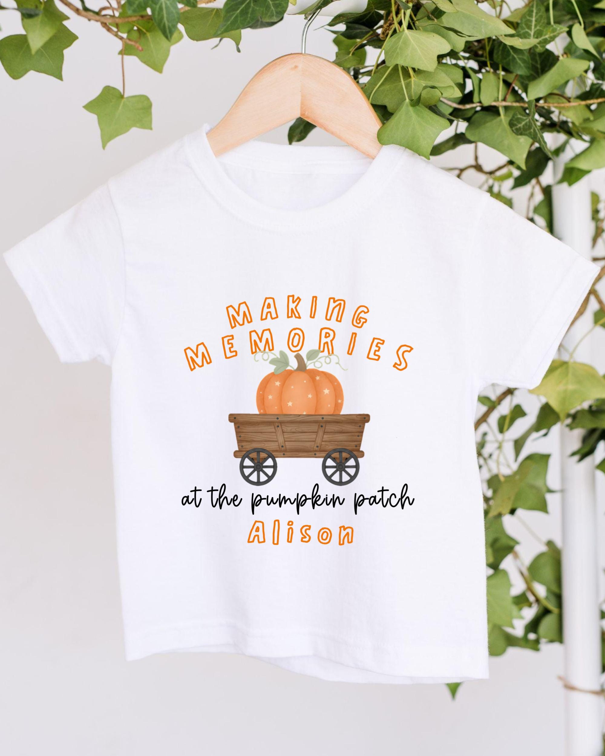 A white t-shirt with a graphic design featuring a pumpkin and a wagon, along with the text "Making memories at the pumpkin patch Alison".