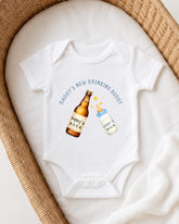 A white onesie with a graphic design on it, featuring a bottle of beer and a baby bottle, along with the text "DADMY'S NEW DRINKING BUDDY".