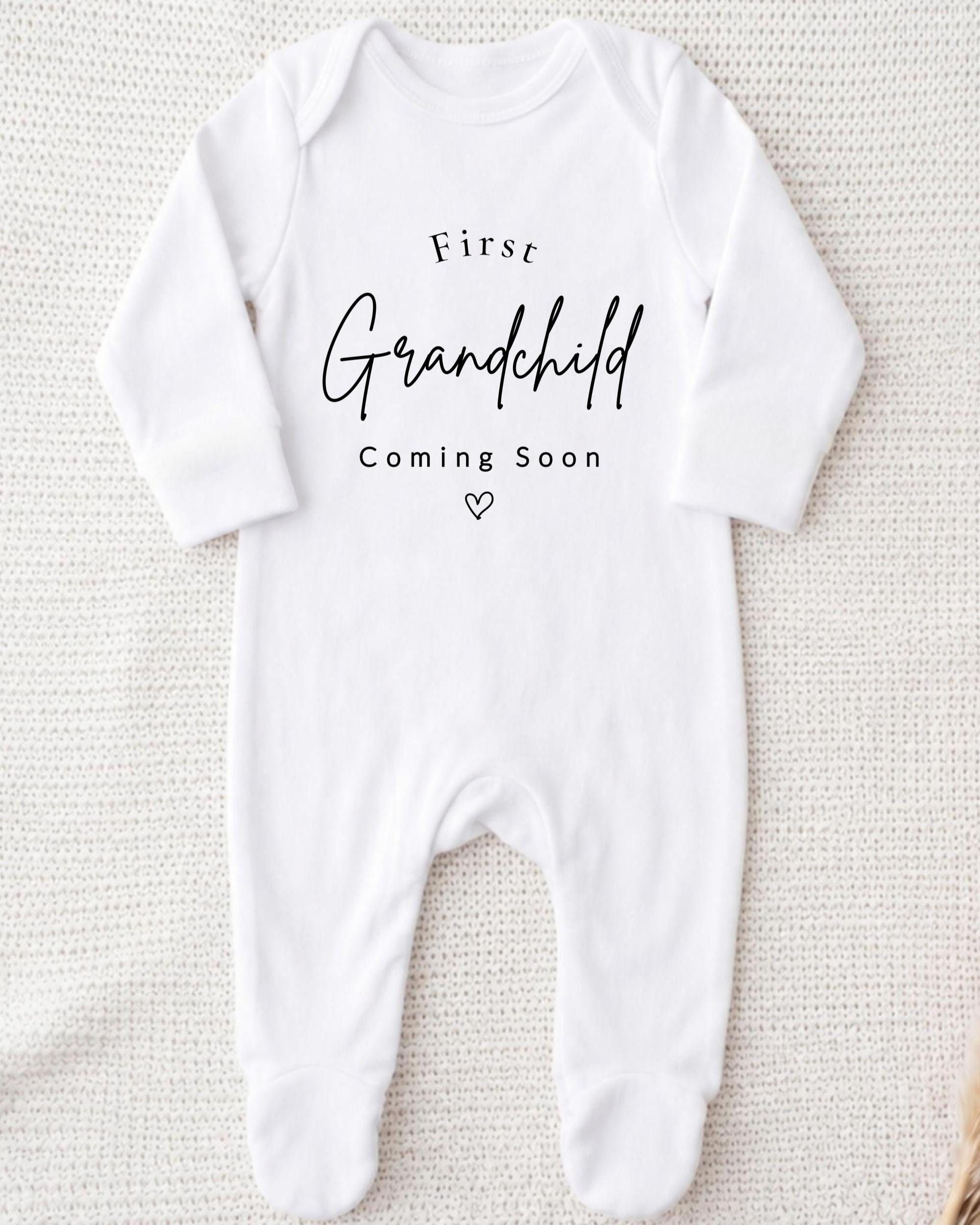 A white onesie with the text "First Grandchild Coming Soon" printed on it.