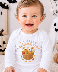 A smiling baby wearing a white onesie with a graphic of a basket of fall fruits and the text "My first Thanksgiving Ollie" printed on it.