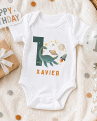 A white onesie with a dinosaur and space-themed design, along with a "XAVIER" name tag, is displayed on a bed with a "Happy Birthday" sign and a balloon.