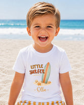 A young boy with a big smile on his face, wearing a white t-shirt with a surfboard graphic and the text "Little Surfer - Ollie" printed on it, standing on a sandy beach with the ocean in the background.