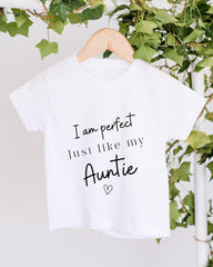 A white t-shirt with black text that reads "I am perfect just like my Auntie" hanging on a wooden hanger.