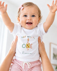 A smiling baby girl wearing a white t-shirt with a birthday message and a colorful candle graphic, being held up by someone's arms.