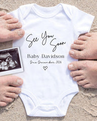 a white baby onesie with the text "See You Soon Baby Davidson Due December 2026" printed on it, surrounded by sand and a black and white photograph of a baby's belly.