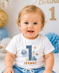 A smiling baby boy wearing a white t-shirt with a graphic design and the name "Johny" printed on it.