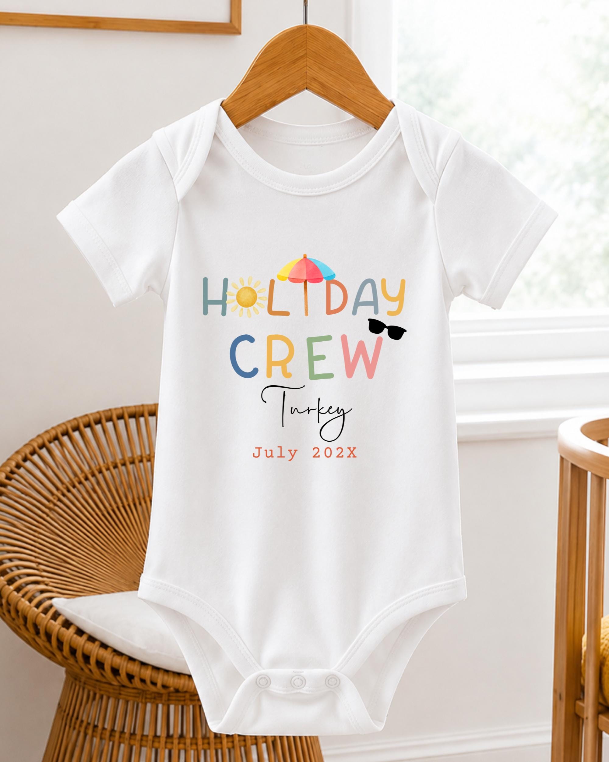 A white baby onesie with the text "HOLIDAY CREW Turkey July 2023" printed on it, hanging on a wooden hanger.