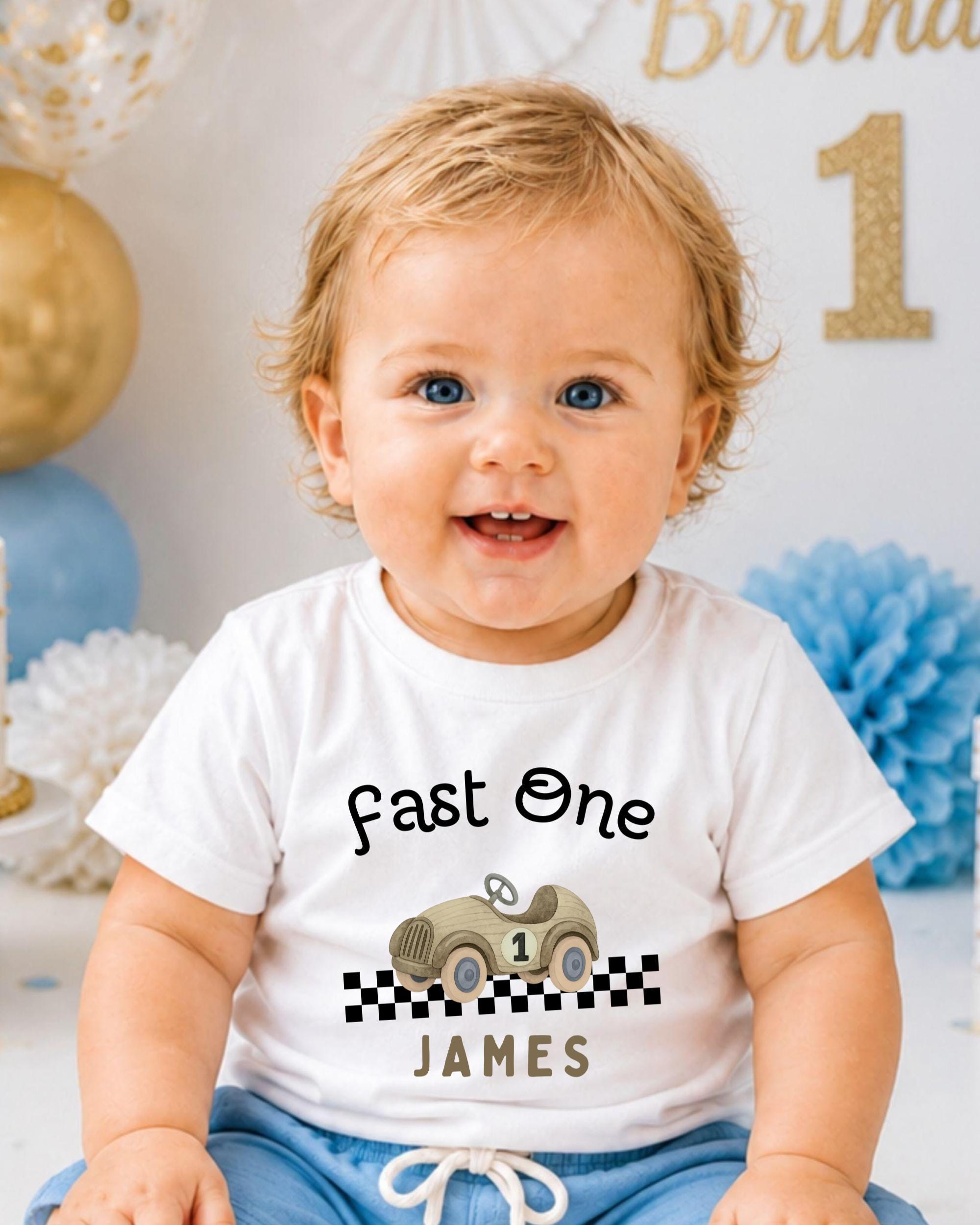 A smiling baby boy wearing a white t-shirt with a race car graphic and the text "Fast One James" is sitting in front of a birthday party backdrop.