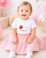 A smiling baby girl wearing a white t-shirt with a strawberry design and a pink skirt, sitting on a pink and white background with balloons and confetti.