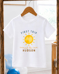 A white t-shirt with a yellow sun graphic and the text "FIRST TRIP around the sun HUDSON" printed on it, hanging on a wooden hanger.