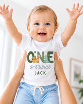 A smiling baby wearing a white shirt with a dinosaur graphic and the text "ONE a-saurus JACK" is being held up by someone's arms.