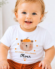 A smiling baby boy wearing a white t-shirt with a cartoon bear and text that says "My first Halloween Oliver 2020".
