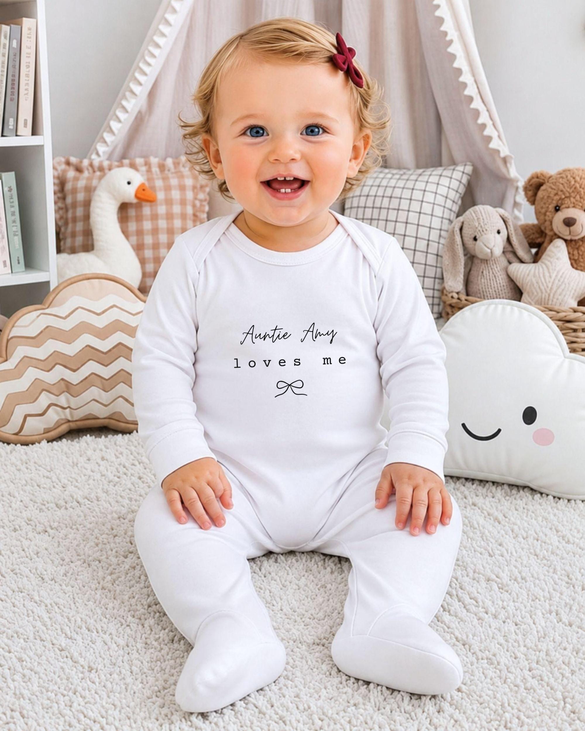 A smiling baby girl wearing a white onesie with the text "Auntie Amy loves me" sits on a white carpet surrounded by stuffed animals.