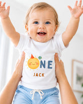 A smiling baby wearing a white shirt with a sun and name printed on it, being held up by someone's arms.