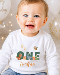 A smiling baby boy wearing a white shirt with the word "One" and a dinosaur graphic, sitting in front of birthday balloons.