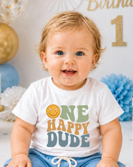 A smiling baby boy wearing a white t-shirt with a happy face and the text "ONE HAPPY DUDE" printed on it.